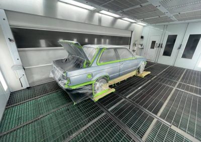Ein silber-blauer BMW Oldtimer steht in einer professionellen Lackierkabine; Fenster, Räder und Kofferraum sind sorgfältig mit Folie und grünem Klebeband für die Lackierung abgeklebt.