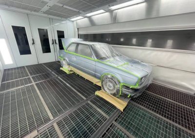 Die Front- und Seitenansicht eines silberfarbenen BMW Oldtimers in einer hellen Lackierkabine. Fenster, Scheinwerfer und Felgen sind sorgfältig mit Klarsichtfolie und neongrünem Klebeband abgeklebt.