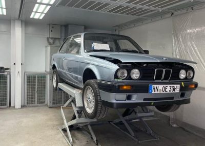 Ein silber-blauer BMW E30 Oldtimer steht auf einer Scherenhebebühne in einer hell beleuchteten Werkstatt für Karosseriearbeiten.