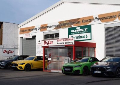 Außenansicht der Dr. Car Werkstatt und Lackiererei in Heilbronn mit mehreren hochwertigen Sportwagen und Premiumfahrzeugen davor, fotografiert aus einer anderen Perspektive.