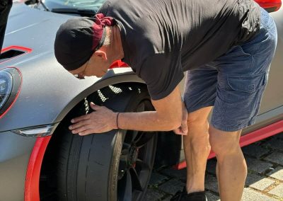 Nahaufnahme unserer Partners, der den vorderen Slick-Reifen eines matt-grauen Porsche 911 GT3 RS mit roten Akzenten prüft, auf dem Nürburgring mit Dr. Car.