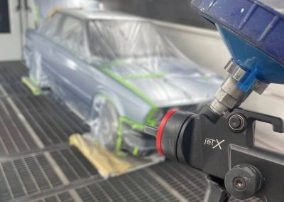 Eine Detailaufnahme einer schwarzen jetX Profi-Lackierpistole mit blauem Farbbecher im Vordergrund. Im unscharfen Hintergrund steht ein vollständig abgeklebter, silberfarbener BMW E30 Oldtimer in einer hellen Lackierkabine auf einem Gitterboden.