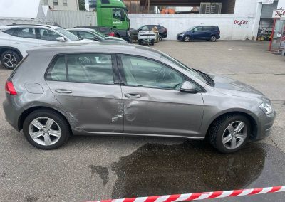 Seitliche Ansicht eines grauen VW Golf mit massivem Unfallschaden an der Tür und dem Kotflügel, bereit zur Karosseriereparatur.